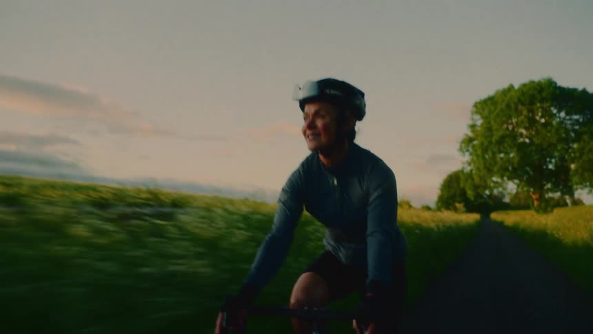 A smiling woman wearing a helmet is riding a bicycle on a path surrounded by bushes and grass fields