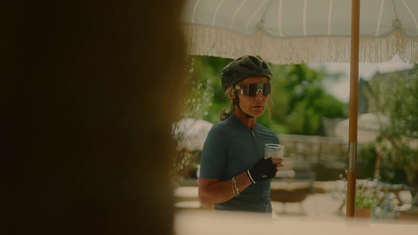 A female cyclist stands under a shade at midday smiling as she drinks water from a cup she is wearing a helmet and cycling glasses