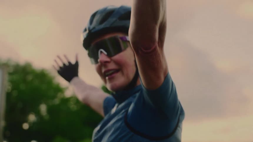 A female cyclist wearing a helmet cycling glasses and sportswear stands outdoors at midday with her arms spread wide in a celebratory pose laughing joyfully