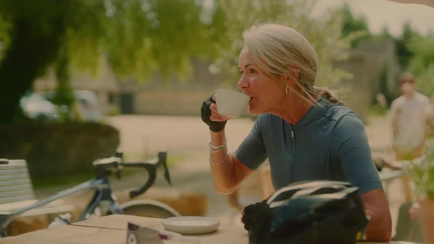 A white haired woman is sitting at an outside table drinking something from a white mug and there is a bicycle helmet on a table