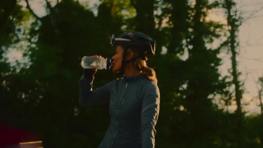A female cyclist on a stationary bicycle near a road happily drinking water from a bottle