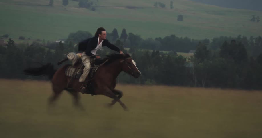 Horseman rides a galloping horse through a field while foggy mountains tower over a town in the background