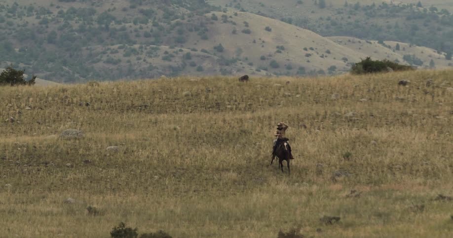 A man wearing a cowboy hat rides a horse across a highland covered in golden plains grasses on a cloudy day