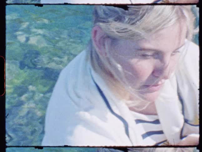 Woman sits by clear blue water in vintagestyle footage wearing light clothing and gazing at the camera