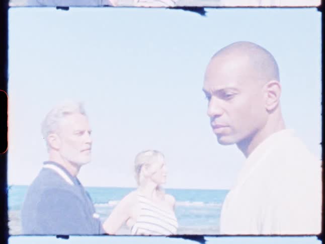 Three people stand on a beach in bright daylight gazing in different directions with thoughtful expressions