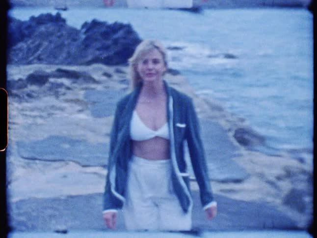 A blonde woman is walking along a rocky ocean coastline