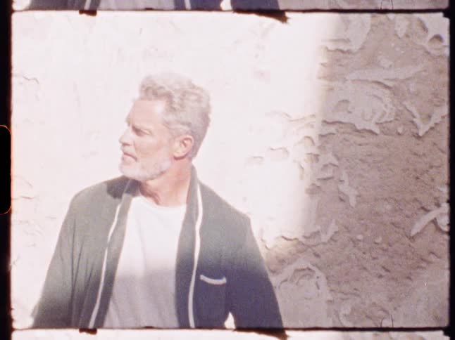 A man wearing a suit stands beside a wall on a sunny midday hands in his pockets with gray beard and hair