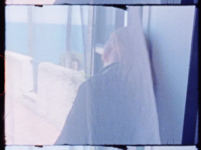 A woman stands at the entrance of a room in a resort as a transparent white curtain billows in the wind at midday