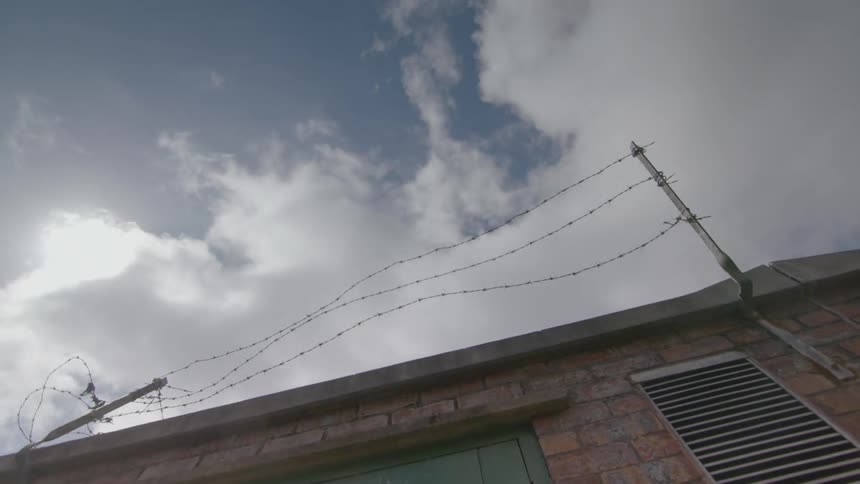 Barbed wire hangs atop a building as clouds pass by