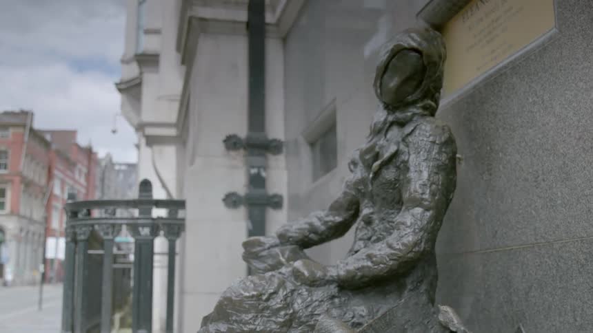 A statue of a woman sitting on a bench sits in front of a building in a city