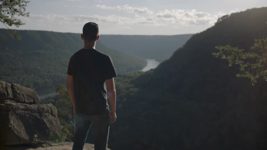 A man stands on a ledge and looks at a view of a river valley