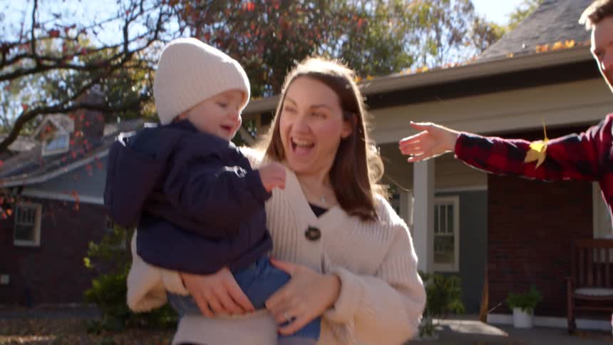 A mother carries her son in the front yard at midday as the father tosses fallen leaves into the air bringing excitement and joy to both the child and the mother