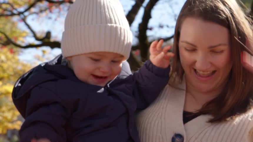 A mother holds her toddler outdoors at midday as the father tosses fallen leaves into the air exciting the baby as the leaves scatter and gently fall to the ground