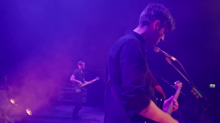 A band performs on a smokefilled stage under purple lighting with multiple musicians in action