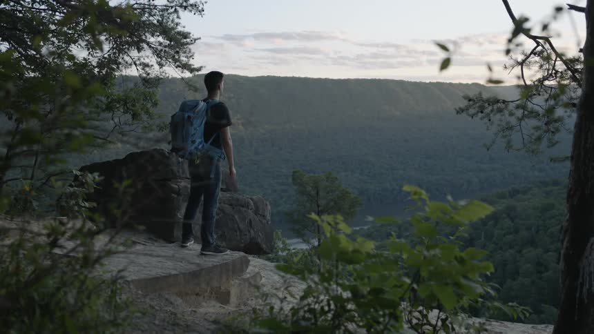 Man wearing a backpack stands at the edge of a cliff overlooking hills