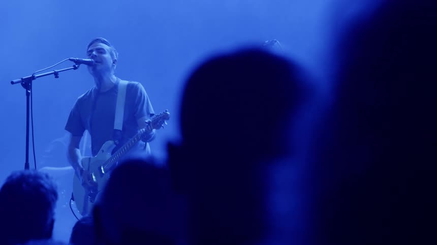 A man plays a guitar and sings into a microphone during a concert performing with a musical band as the crowd watches attentively