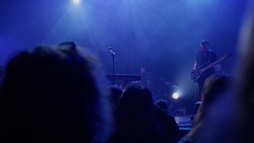 A musician plays bass guitar on a bluelit stage with a microphone stand centered under overhead lights