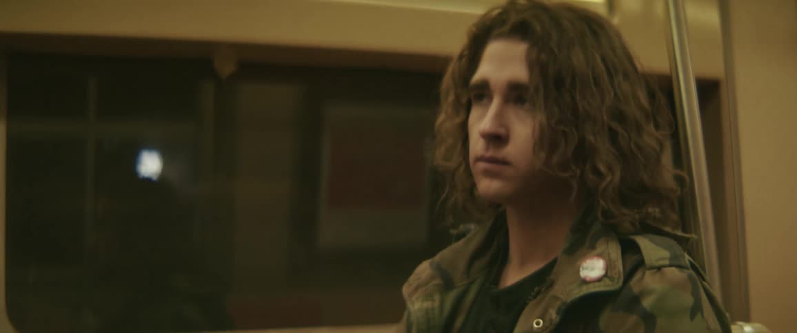 A young man wearing a camouflage jacket stands during a train ride at night checking his watch for the time