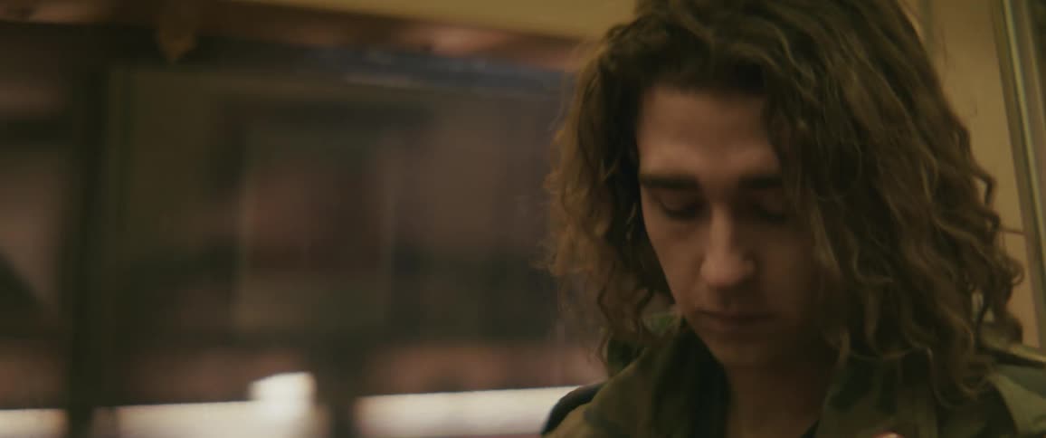 A young man with long curly hair in a camouflage jacket sits in a moving subway car glancing at his wristwatch with a sad impatient expression