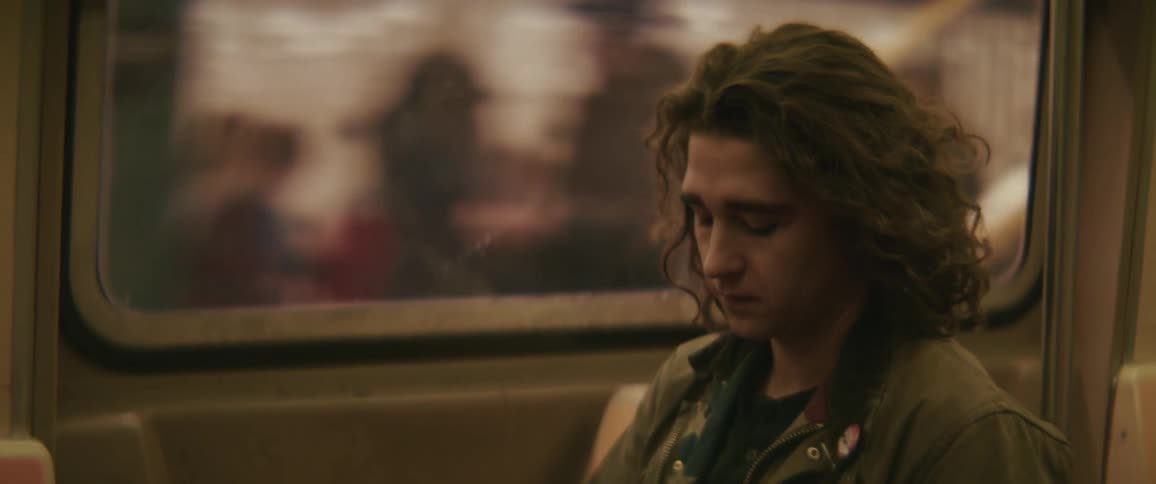 The face of a man with long hair who is sitting on a train near the window and he is putting something in his ear