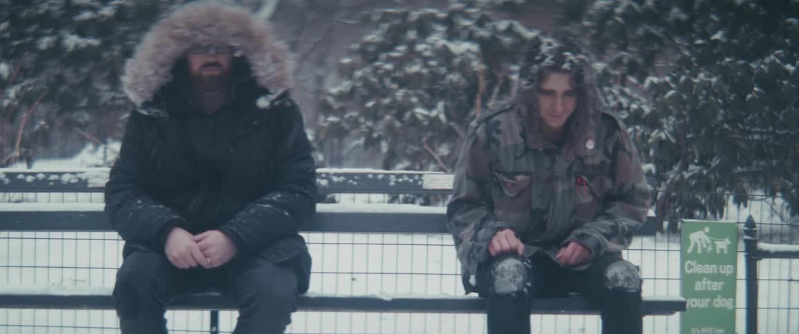 A man walks up to a homeless man sitting on a bench beside the street shivering from the cold and snow the man wearing a jacket hands an object to the homeless man and then walks away at midday