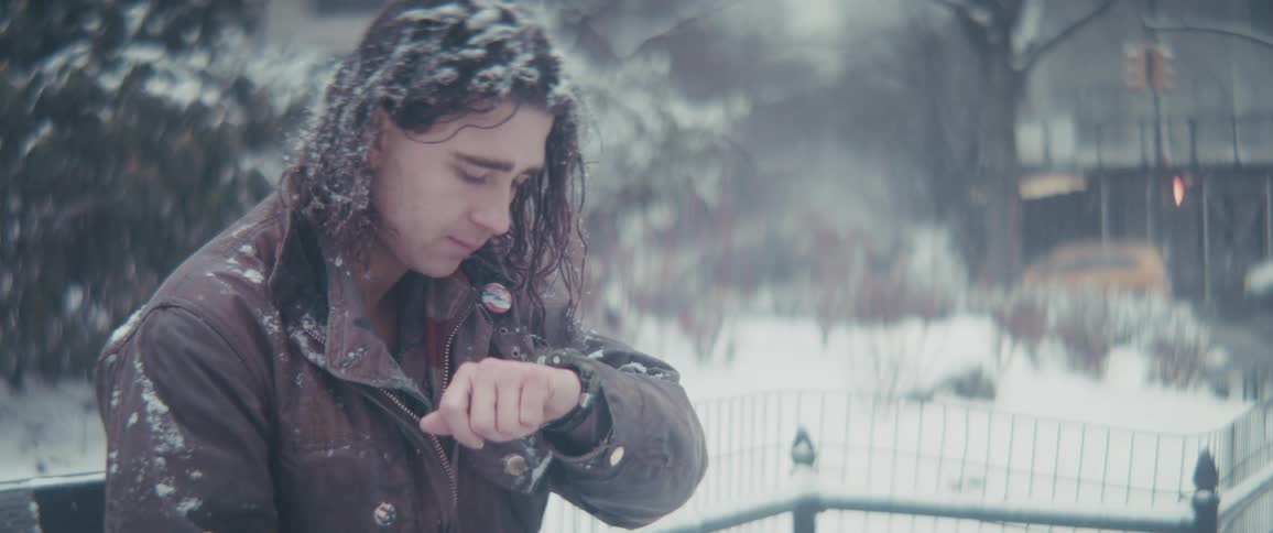 The face of a man with long hair wearing a brown coat who is sitting outside and is being hit by a blizzard