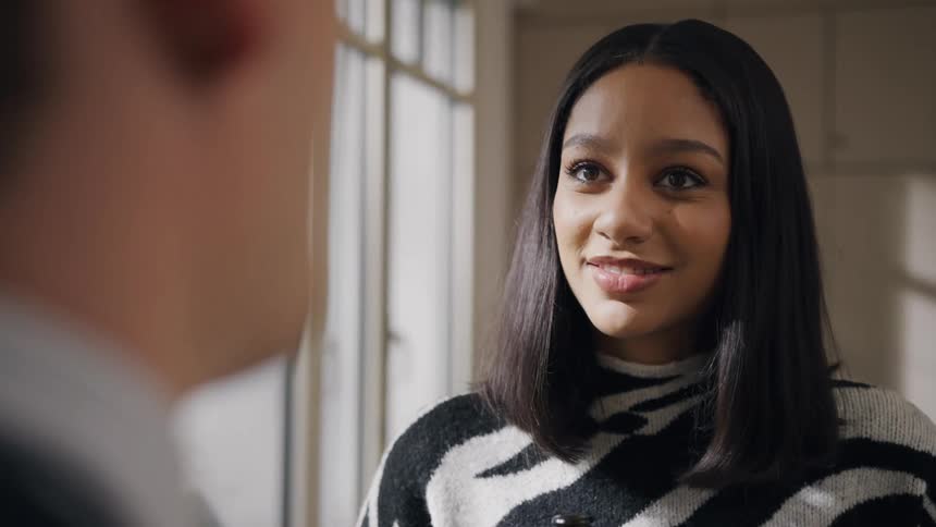 A young black woman talks to a man as they both stand beside a window at midday