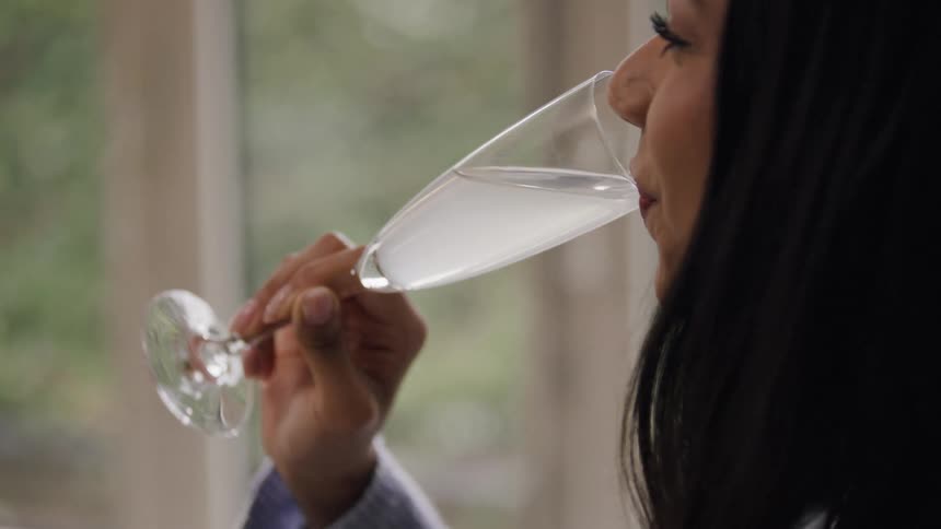A woman sips from a champagne flute near a bright window