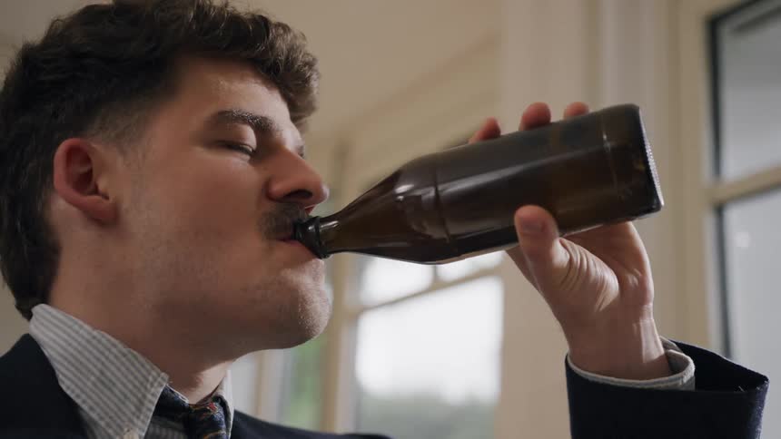 A man drinks from a brown glass bottle indoors