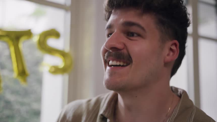 A group of friends greets each other by a window framed with gold foil balloons