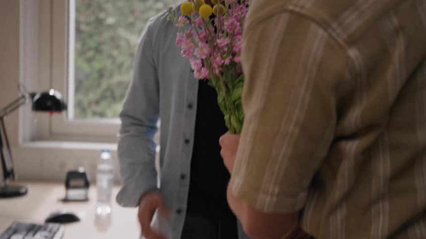 A man visits his friend in his office workspace at midday gives him a hug and presents a flower to congratulate him