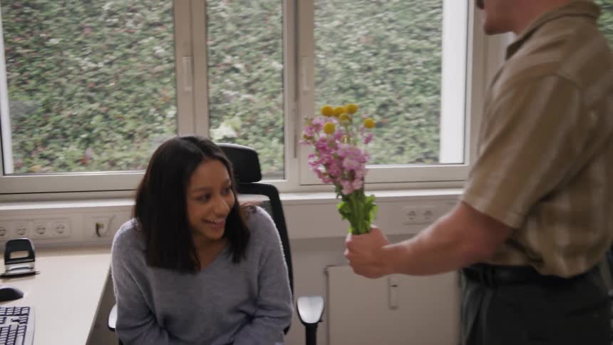 A man walks up to a coworker and gives her flowers