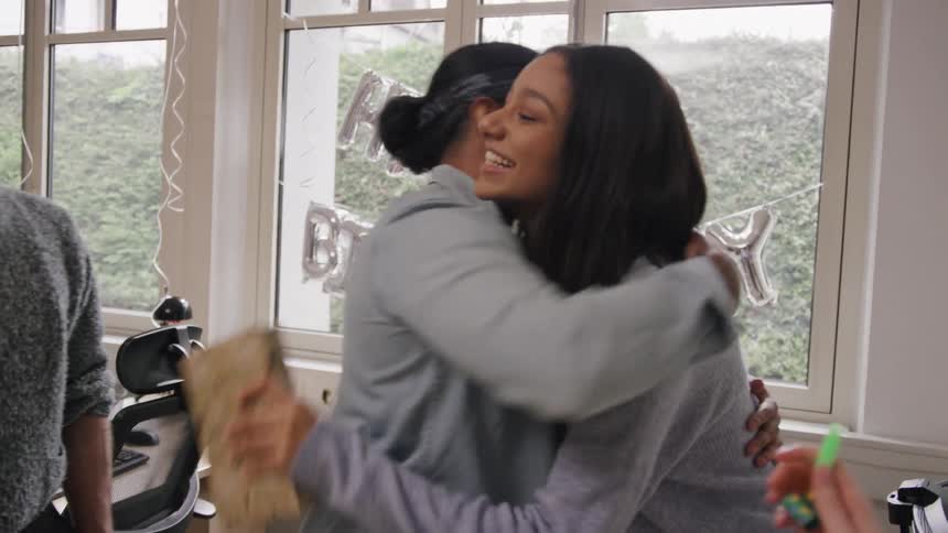 An office party takes place at midday as a worker is surprised with a birthday celebration with female coworkers hugging him and handing him a present