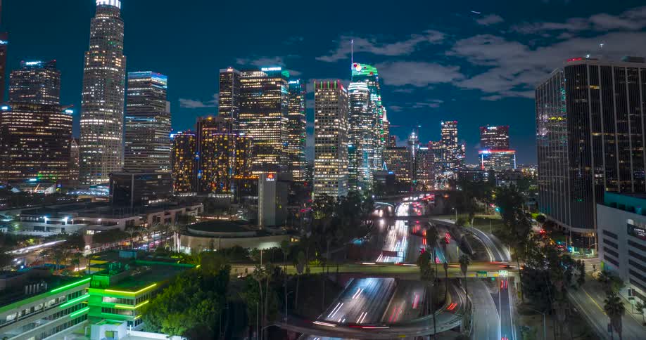 A vibrant aerial timelapse captures the Los Angeles skyline at night with glowing skyscrapers rising behind the light trails of traffic on a busy freeway