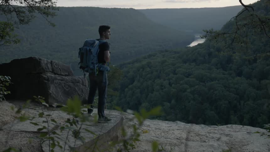 Man wearing a backpack stands at the edge of a cliff overlooking hills