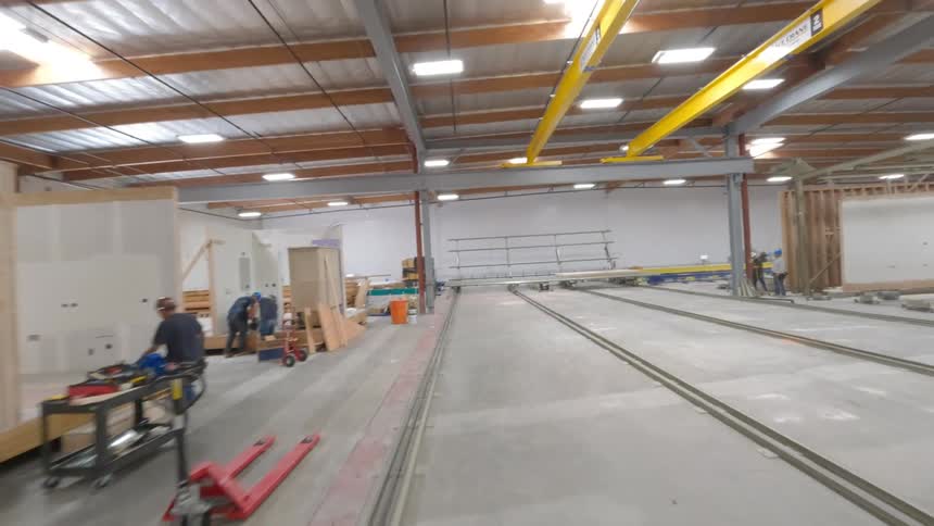 An FPV drone flies through a large manufacturing warehouse capturing workers and machinery on an assembly line building prefabricated housing components