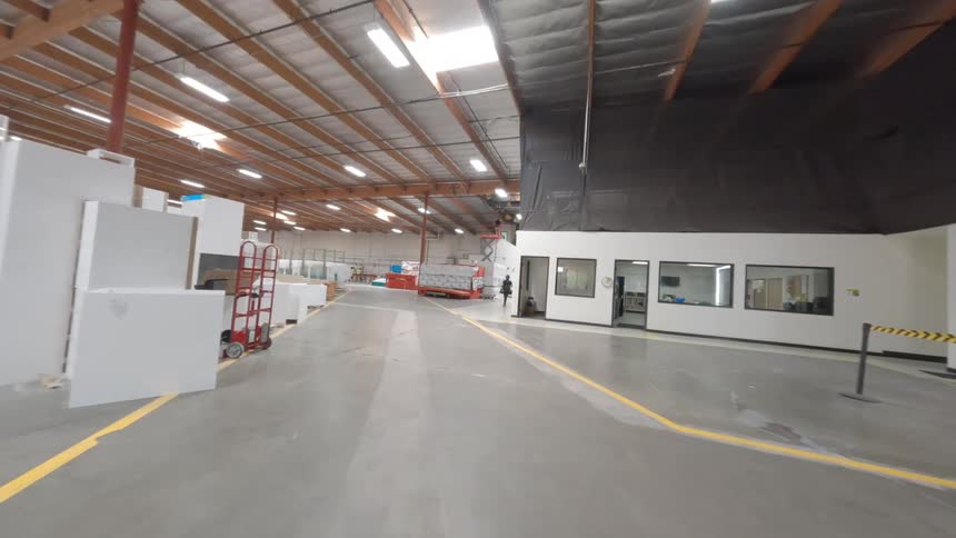 An FPV drone captures a wide view of a busy warehouse interior showing forklifts in motion various industrial machinery stacks of lumber and workers moving throughout the space