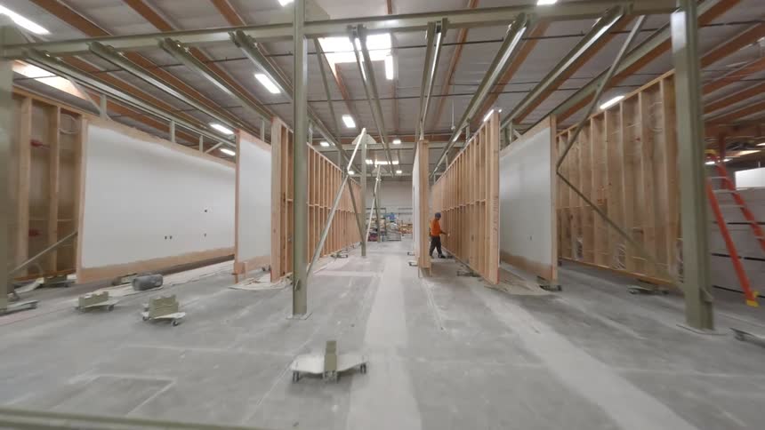 A FPV drone flying inside a  factory warehouse filled with wood and lumber with workers wearing green vests