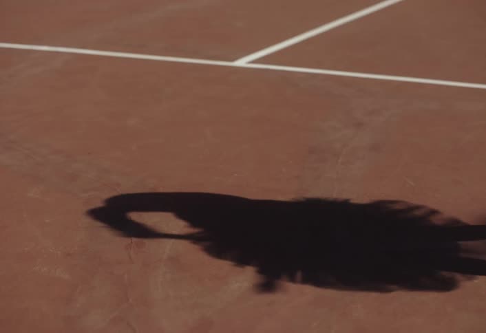 The silhouette of a person playing tennis using a racket on a red court with white lines