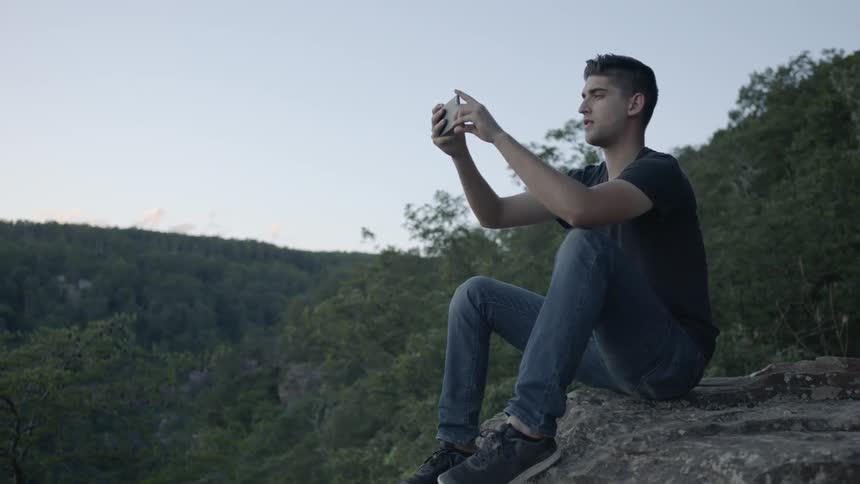 A man takes a photo while sitting on the edge of a cliff