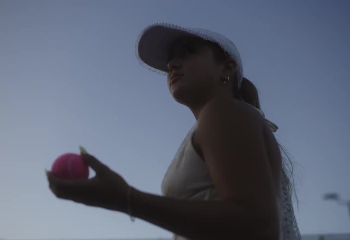 A young woman wearing a baseball hat holds a tennis ball tossing it up with her hands at sundown