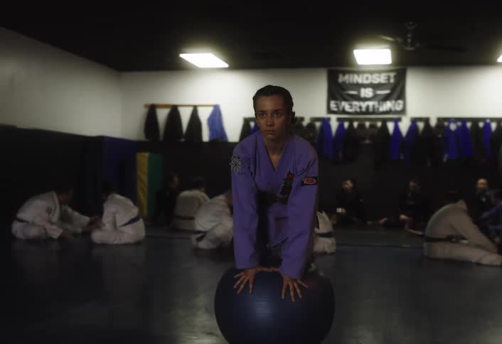 Female judoka leaning on a ball in a gym and other students in the background