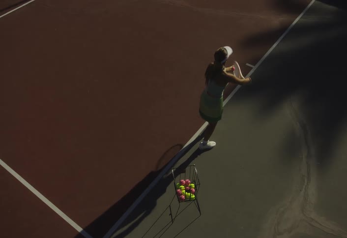 A woman plays on a tennis court with a racket near a basket of balls under dramatic lighting