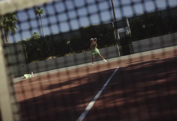 A woman plays tennis on a clay court viewed through the net