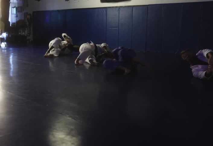 Several individuals in white gis are practicing grappling techniques on a padded mat under low lighting