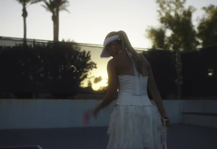 Woman bounces a tennis ball and serves while playing tennis in an outdoor court at sunset
