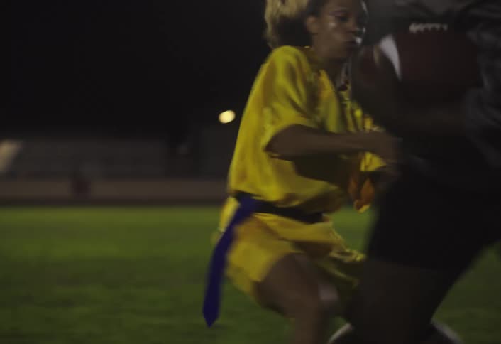 A woman wearing a yellow jersey tackles a man during a football match snatching the ball from him at night