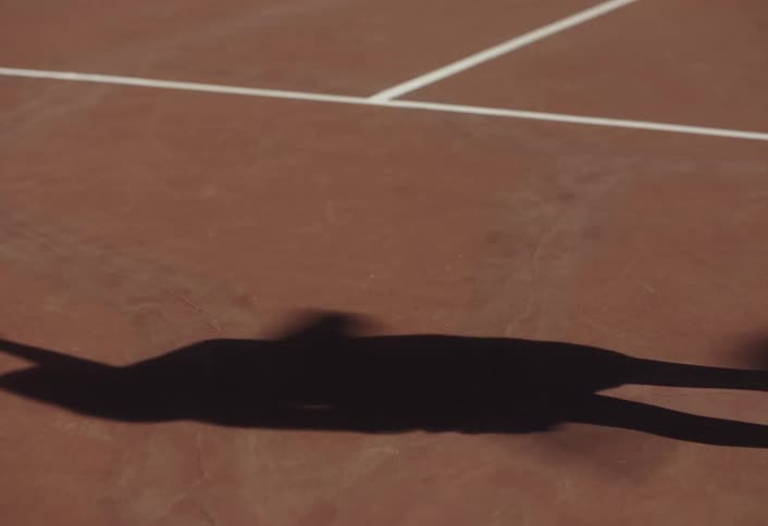 The shadow of a female tennis player serving a tennis ball overhand on a red tennis court during the day