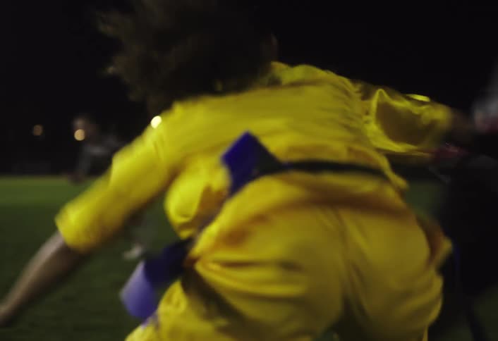 Two women play rugby on a grassy field at nightone sprints in dark athletic gear while the other in a yellow top moves to block her