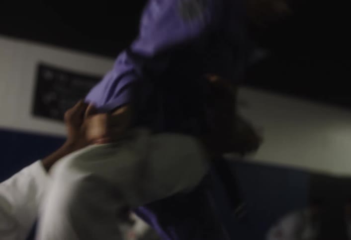 A group of students wearing martial arts uniforms engage in a grappling hold on a dark floor